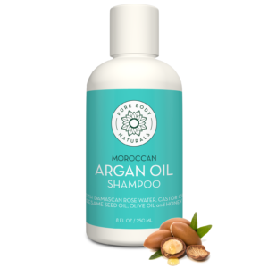 A bottle of Argan Oil Hydrating Shampoo, for All Hair Types with a light teal label and white cap. The label mentions it contains Damascus rose water, castor oil, sesame seed oil, olive oil, and honey. Some argan nuts and green leaves are placed in front of the bottle.